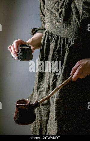 Femme en robe sombre tenir dans les mains artisanat céramique pot de café cezve avec une longue poignée en bois et un pot de lait pour faire du café turc. Banque D'Images