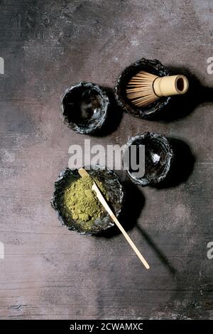 Poudre de thé vert matcha japonais dans un bol en céramique artisanale, deux tasses, fouet en bambou sur fond de texture sombre. Pose à plat, espace Banque D'Images