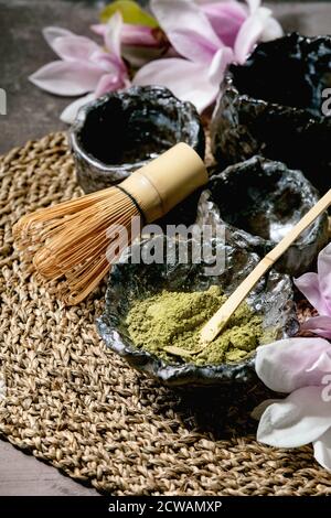 Poudre de thé vert matcha japonais dans un bol en céramique artisanale, deux tasses, fouet en bambou et fleurs de magnolia rose sur une serviette en paille sur fond de texture sombre Banque D'Images