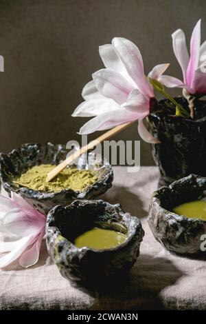 Thé vert de matcha japonais dans des tasses en céramique artisanale avec poudre de matcha, fouet en bambou et fleurs de magnolia rose sur toile de table grise. Banque D'Images