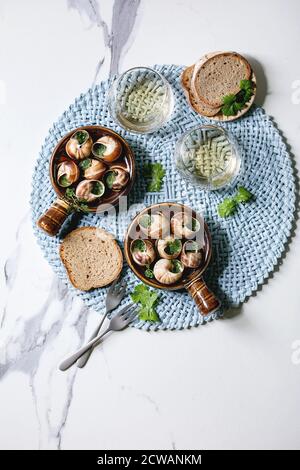 Escargots de Bourgogne - escargots au beurre d'herbes, plat gastronomique, dans une casserole traditionnelle en céramique avec coriandre, pain, verre de vin blanc sur serviette bleue Banque D'Images