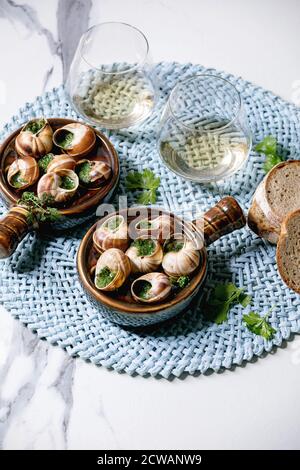 Escargots de Bourgogne - escargots au beurre d'herbes, plat gastronomique, dans une casserole traditionnelle en céramique avec coriandre, pain, verre de vin blanc sur serviette bleue Banque D'Images