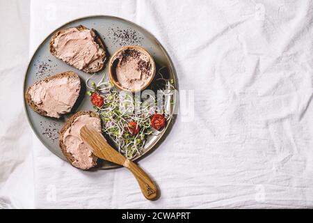 Sandwiches et bol en céramique de pâté de foie de poulet maison avec couteau en bois, tomates séchées et salade verte sur plaque grise sur lin blanc Banque D'Images