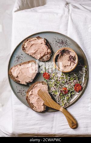 Sandwiches et bol en céramique de pâté de foie de poulet maison avec couteau en bois, tomates séchées et salade verte sur plaque grise sur lin blanc Banque D'Images