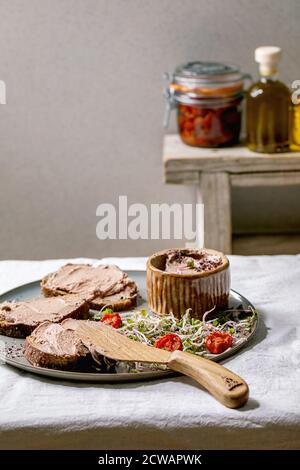 Sandwiches et bol en céramique de pâté de foie de poulet maison avec couteau en bois, tomates séchées et salade verte sur plaque grise sur lin blanc Banque D'Images