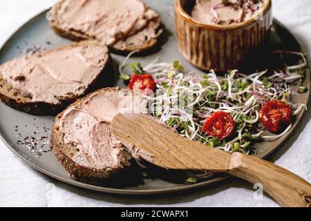 Sandwiches et bol en céramique de pâté de foie de poulet maison avec couteau en bois, tomates séchées et salade verte sur plaque grise sur lin blanc Banque D'Images