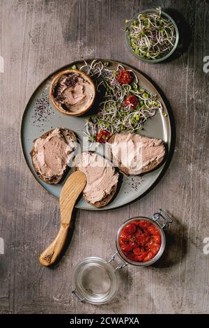 Sandwiches et bol en céramique de pâté de foie de poulet maison avec couteau en bois, tomates séchées et salade verte sur assiette grise sur texture sombre Banque D'Images