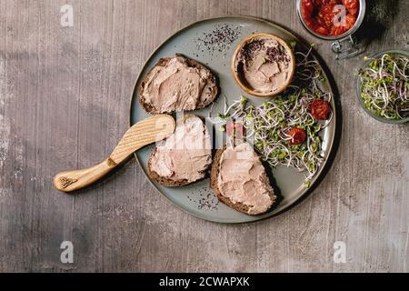 Sandwiches et bol en céramique de pâté de foie de poulet maison avec couteau en bois, tomates séchées et salade verte sur assiette grise sur texture sombre Banque D'Images