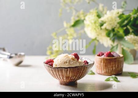 Glace à la vanille au caramel maison avec framboises congelées dans un bol en céramique placé sur une table en marbre blanc avec fleurs derrière. Banque D'Images