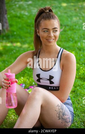 Une jeune femme portant des vêtements de sport assise dehors sur l'herbe tenant une bouteille d'eau rose. Banque D'Images
