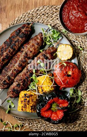 Restauration rapide de l'est. Rôti de boeuf épicé, lyulya kebab sur des bâtonnets sur l'assiette avec légumes grillés, épis de maïs doux, tomate et paprika, sauce tomate sur bois Banque D'Images