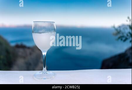 Gros plan d'un verre à tige d'ouzo sur un blanc Admirez le coucher du soleil avec la caldeira de Santorin et la mer bleue en arrière-plan Banque D'Images