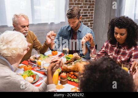 foyer sélectif de la famille multiethnique avec les yeux fermés tenant les mains pendant le dîner le jour de thanksgiving Banque D'Images