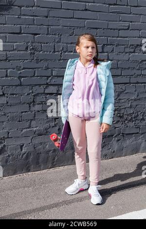 Petite fille en vêtements colorés avec un support de skateboard près du mur de brique gris, photo verticale Banque D'Images