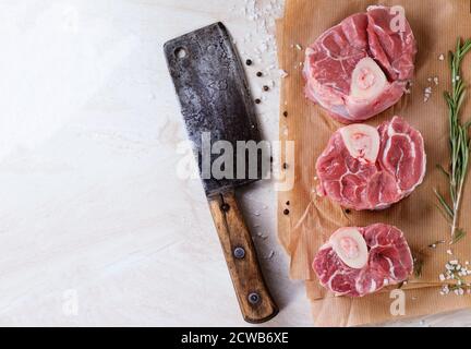 Viande Osso-buco crue sur papier froissé avec sel, poivre et romarin. Avec une épée rétro sur du marbre blanc comme arrière-plan. Vue de dessus. Banque D'Images