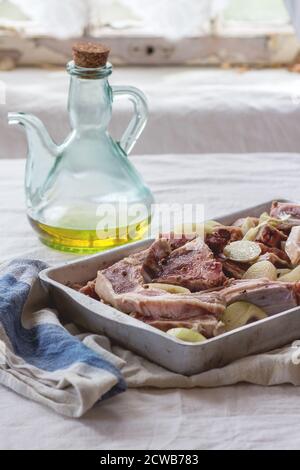 Mariner des côtelettes d'agneau crues avec de l'oignon, de l'huile d'olive et du poivre dans un plat à pâtisserie en aluminium sur une table de cuisine avec nappe blanche. Lumière naturelle. Banque D'Images