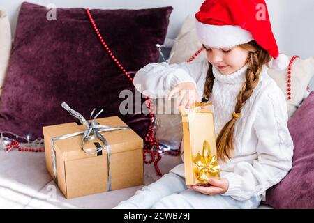 Une fille dans un chapeau de père noël est assise sur le canapé et ouvre un cadeau de nouvel an dans un or package Banque D'Images