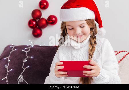 Une petite fille dans un chandail blanc et un Père Noël Le chapeau Claus regarde un cadeau dans une boîte rouge et se réjouit Banque D'Images