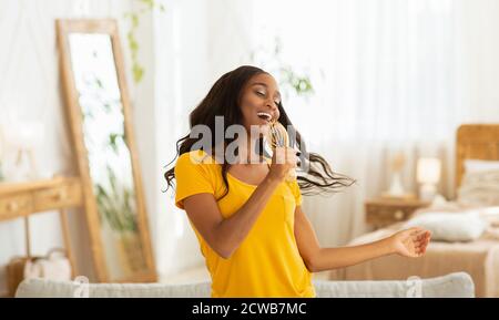 Joyeuse femme noire avec brosse à cheveux comme le chant et la danse de microphone à la maison Banque D'Images