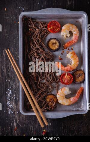 Dîner asiatique avec nouilles soba, champignons shiitake, tomates grillées et crevettes dans un plat à pâtisserie en aluminium avec baguettes en bois sur du vieux bois Banque D'Images