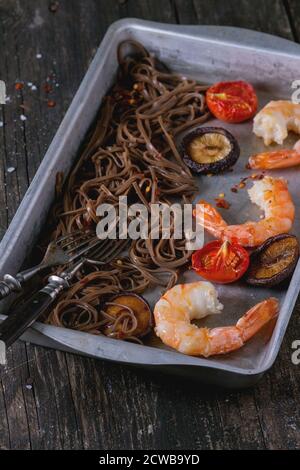 Dîner de style asiatique avec des nouilles soba, des champignons shiitake, des tomates grillées et les crevettes dans un plat de cuisson en aluminium avec plus de fourches vintage ancienne en bois retour Banque D'Images