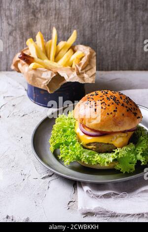 Hamburger frais maison aux graines de sésame noir dans une assiette métallique vintage avec pommes de terre frites, servi sur une table à plâtres blanche avec de la ba en bois gris Banque D'Images