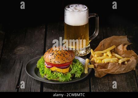 Hamburger maison frais avec dans une ancienne plaque métallique avec pommes de terre frites en papier de cuisson, servi avec un verre de bière froide sur une ancienne table en bois W Banque D'Images