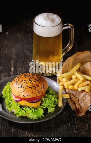 Hamburger maison frais avec dans une ancienne plaque métallique avec pommes de terre frites en papier de cuisson, servi avec un verre de bière froide sur une ancienne table en bois avec dar Banque D'Images