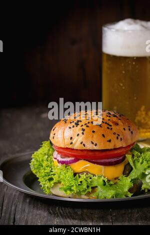 Hamburger maison frais avec dans une ancienne plaque métallique avec pommes de terre frites en papier de cuisson, servi avec un verre de bière froide sur une ancienne table en bois avec dar Banque D'Images