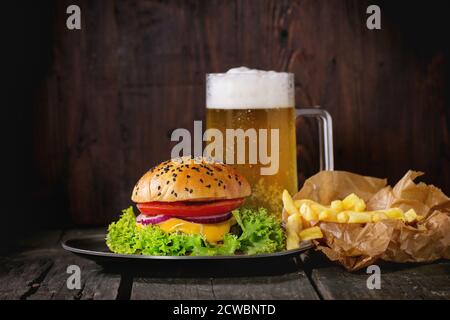 Hamburger maison frais avec dans une ancienne plaque métallique avec pommes de terre frites en papier de cuisson, servi avec un verre de bière froide sur une ancienne table en bois avec dar Banque D'Images