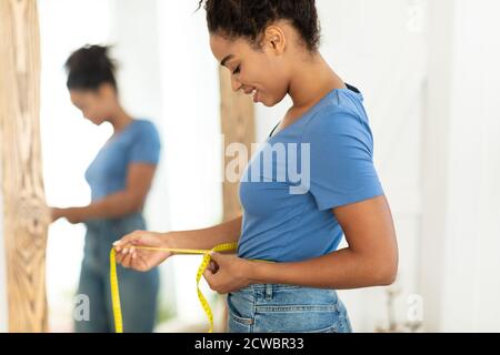 Femme africaine souriante mesurant la taille après avoir fait un slimming debout à la maison Banque D'Images