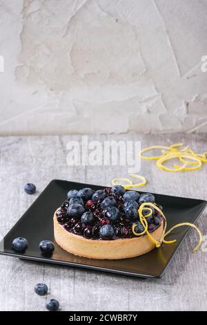 Tarte au citron ronde avec des bleuets frais et cuits, servis sur plaque carrée noire avec les zestes de citron sur fond de bois gris. Banque D'Images