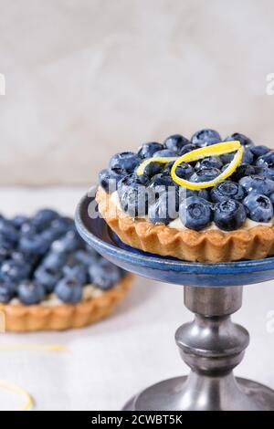 Tartelette au citron aux myrtilles fraîches et zeste de citron, servie sur un support à gâteau vintage sur une nappe blanche. Banque D'Images