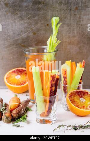 Émincé de carottes et de céleri crus et colorés comme en-cas végétarien, d'oranges sanguines et de thym avec une tasse de verre d'orange frais et de jus de carottes sur la pierre blanche Banque D'Images