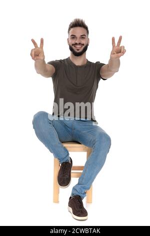 Positif homme décontracté gestant la paix avec les deux mains et souriant assis sur une chaise sur fond blanc de studio Banque D'Images