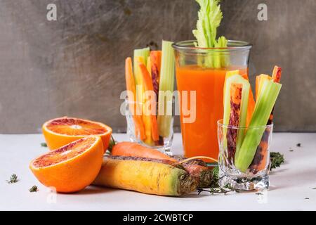 Émincé de carottes et de céleri crus et colorés comme en-cas végétarien, d'oranges sanguines et de thym avec une tasse de verre d'orange frais et de jus de carottes sur la pierre blanche Banque D'Images