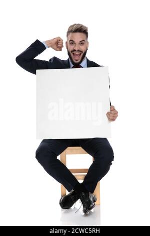 un jeune homme d'affaires heureux tenant une planche vide et célébrant la victoire, tenant le poing dans les airs, assis isolé sur fond blanc Banque D'Images