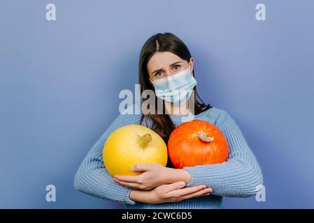 Une jeune femme malheureuse et fatiguée porte un masque médical, tient deux petites citrouilles, a une expression faciale insatisfaite, vêtue d'un pull tricoté Banque D'Images