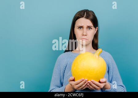 Une jeune femme malheureuse et fatiguée se sent dégoût, tient une petite citrouille jaune mûre, a une expression faciale insatisfaite, vêtue d'un pull en maille Banque D'Images