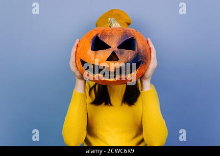 Jeune femme méconnaissable isolée sur fond de studio violet qui cache son visage derrière une grosse citrouille sombre sculptée, vêtue d'un chandail et d'un chapeau orange Banque D'Images