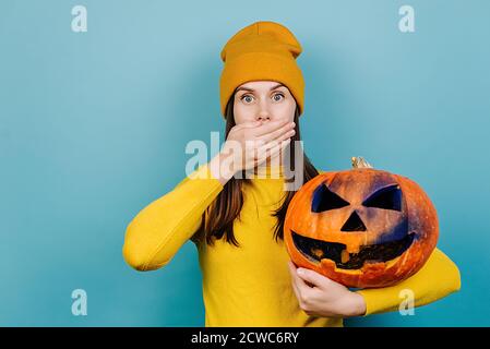 Une jeune femme stupéfiée tenant une grosse citrouille sculptée effrayante couvrant la bouche avec la main, regardant dans le choc à l'appareil photo, vêtue d'un chandail orange et d'un chapeau Banque D'Images