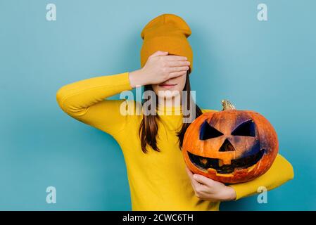 Portrait d'une jeune femme caucasienne qui ferme les yeux avec la main, tenant une grosse citrouille sculptée effrayante, vêtue d'un chandail orange et d'un chapeau Banque D'Images