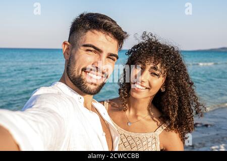 Beau couple de course mixte prenant un selfie à la mer Au crépuscule - beau jeune homme en chemise blanche ouverte avec sa petite amie à poil dur latina sourit Banque D'Images