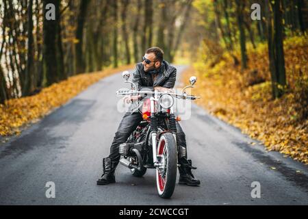 Homme brutal barbu en lunettes de soleil et veste en cuir assis sur une moto sur la route dans la forêt avec un fond coloré et coloré. Banque D'Images