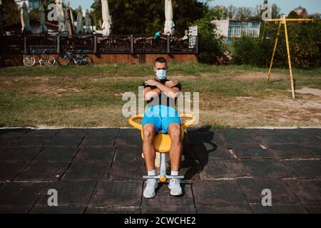 Beau jeune athlète de race blanche avec masque facial s'entraîne sur l'équipement des bras et des jambes dans une salle de sport en plein air. Athlète musclé en plein air. COVID - 19 Banque D'Images