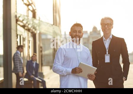 Portrait de partenaires d'affaires multiethniques souriant à l'appareil-photo tout en étant debout ensemble à l'extérieur avec leurs collègues en arrière-plan Banque D'Images