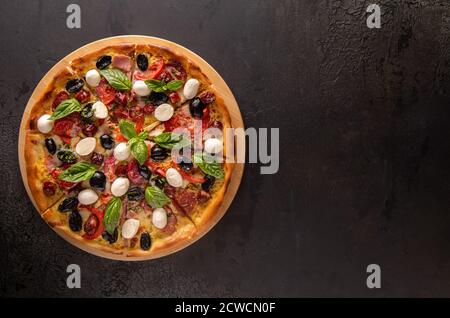 Pizza italienne avec jambon, salami, olives noires, mozzarella, tomates rouges et feuilles de basilic sur plateau de coupe rond en bambou et sur texture noire sur Banque D'Images