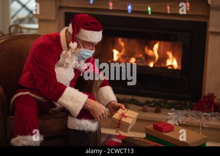 Homme caucasien senior à la maison habillé comme Père Noël, portant un masque de visage, assis sur une chaise près de la cheminée, donnant des cadeaux. Distance sociale pendant la durée Banque D'Images