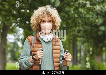 Portrait d'une jeune femme avec des cheveux bouclés portant un masque de protection regarder la caméra pendant la marche sportive dans le parc Banque D'Images