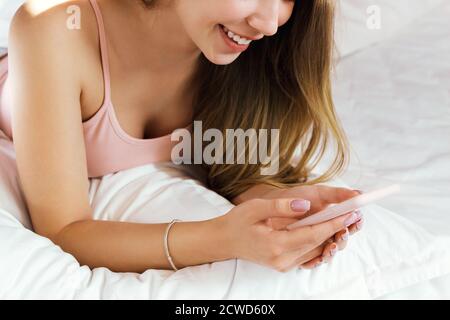 Jeune femme souriante couchée dans un lit blanc et utilisant un téléphone dans sa chambre. Bonjour. Banque D'Images
